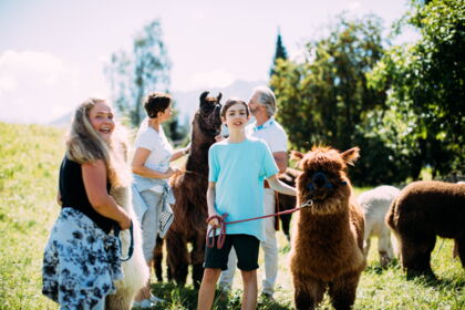 Familien genießen Alpakaspaziergang in sonniger Tiroler Landschaft mit glücklichen Kindern.