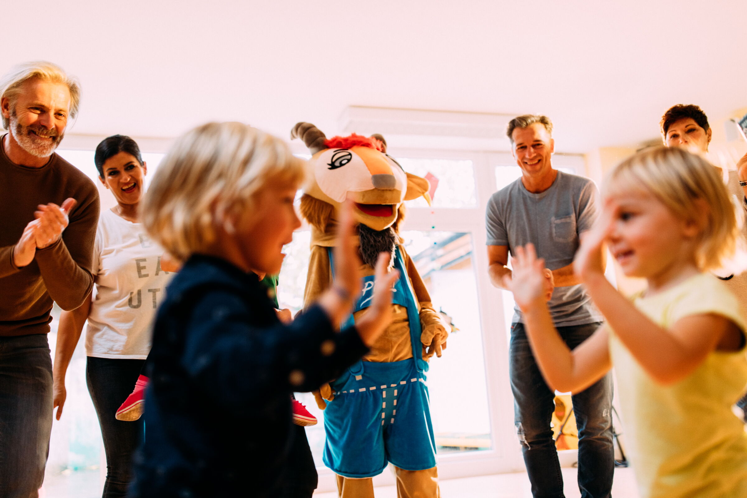 Kinder tanzen und lachen mit Maskottchen und Erwachsenen in hellem Raum.