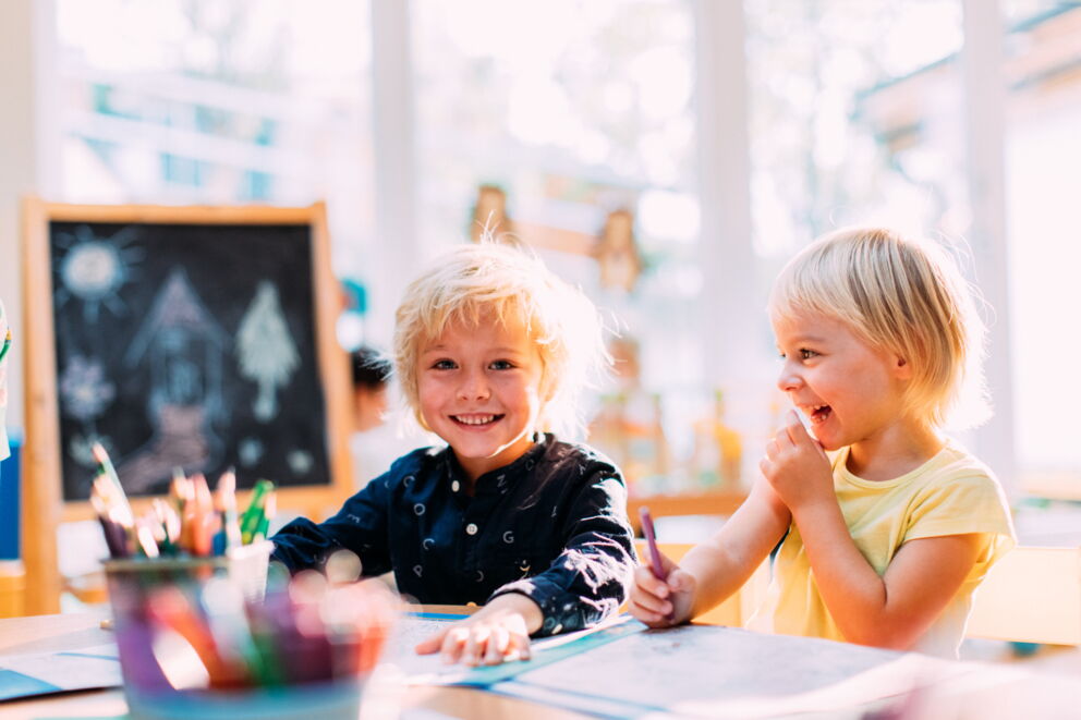 Zwei fröhliche Kinder malen gemeinsam am Tisch in einem hellen Raum.