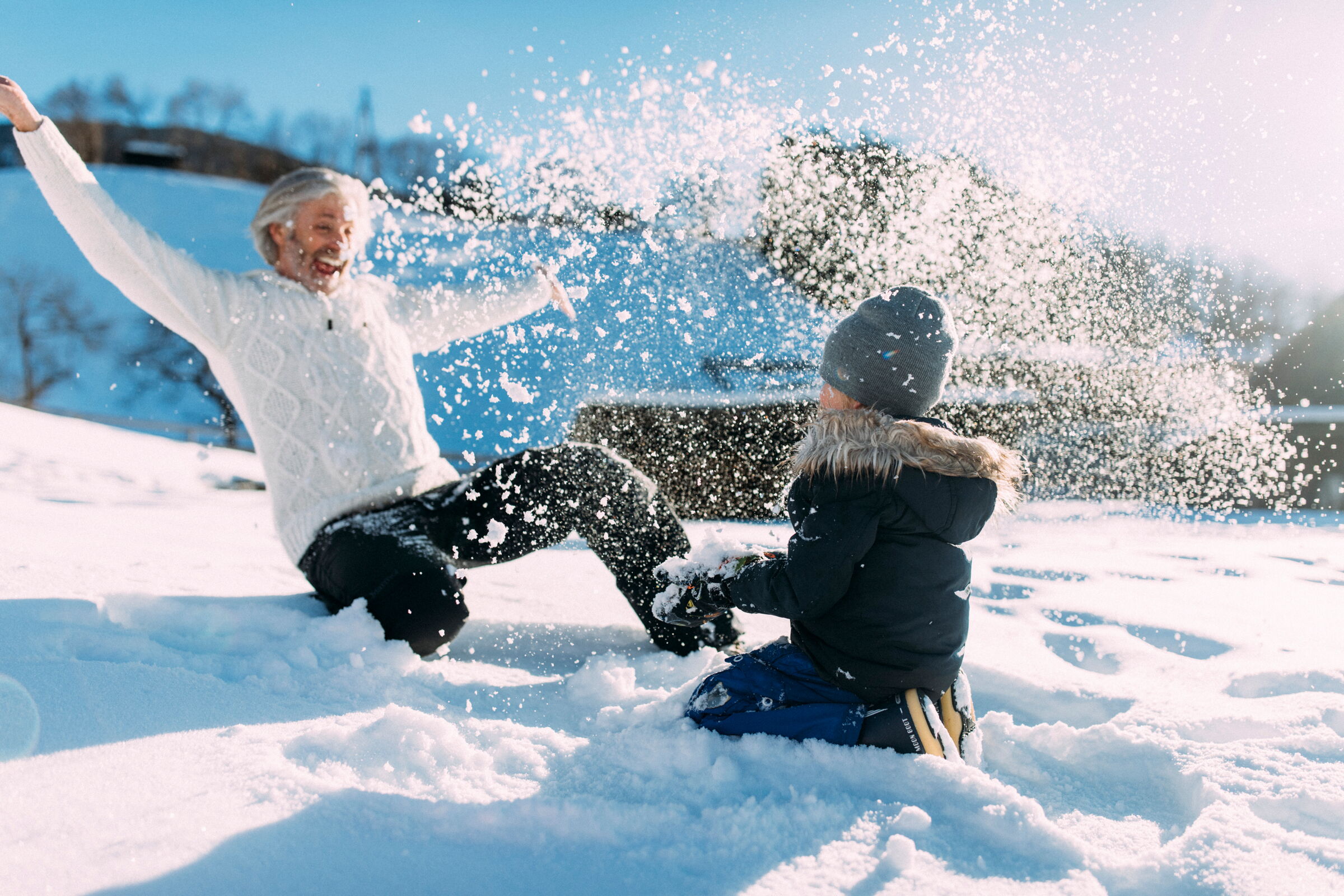 Erwachsene und Kind lachen und spielen im glitzernden Schnee unter strahlend blauem Himmel.