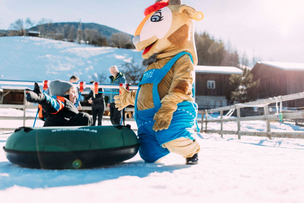 Kind im Schneetubing mit Maskottchen im sonnigen Winterlandschaft.