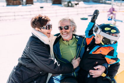 Familie lacht im Winterurlaub mit Schnee im Hintergrund, alle tragen warme Kleidung.
