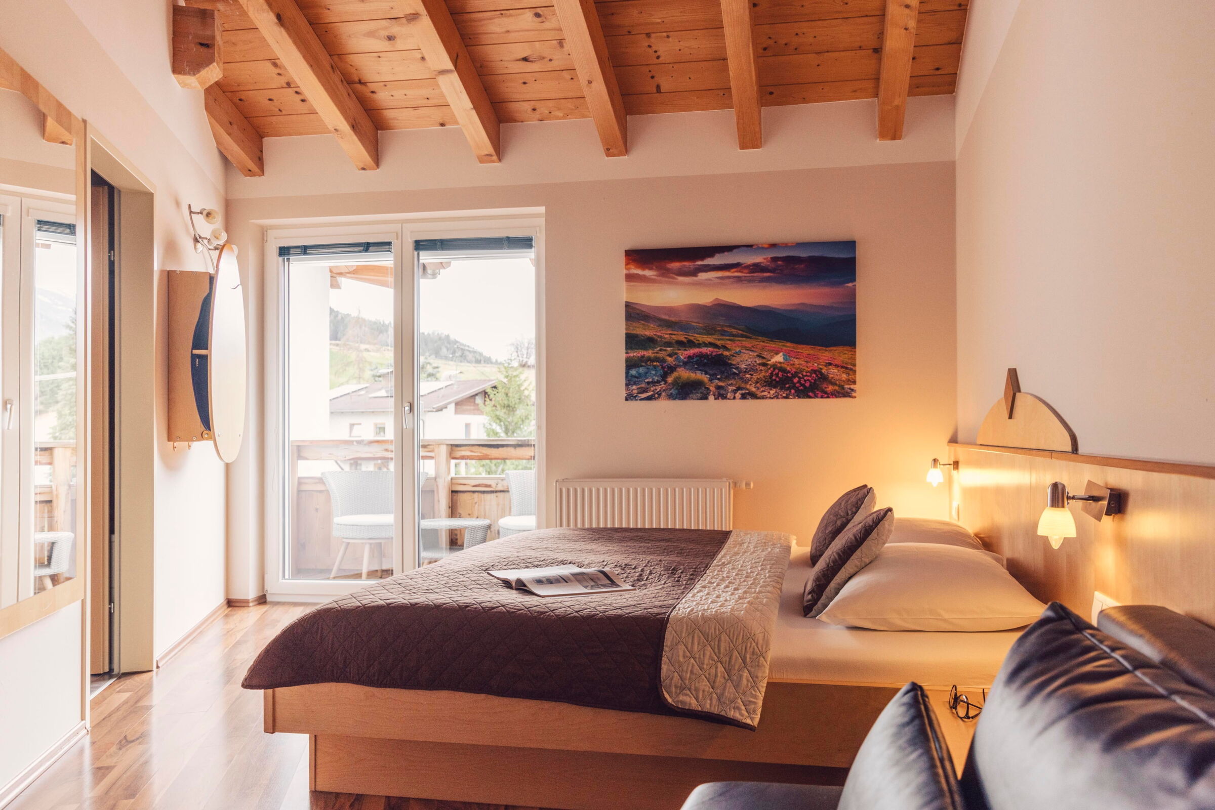 Gemütliches Zimmer mit Holzbalkendecke, großem Bett, Balkon und Bergblick bei Sonnenuntergang.