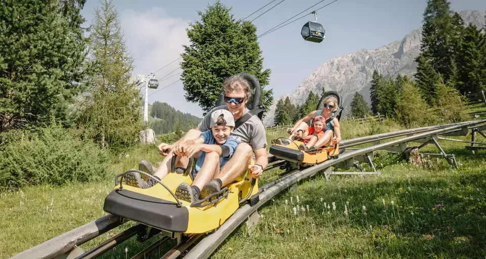 A family rides together on two separate cars on a toboggan run in summer.