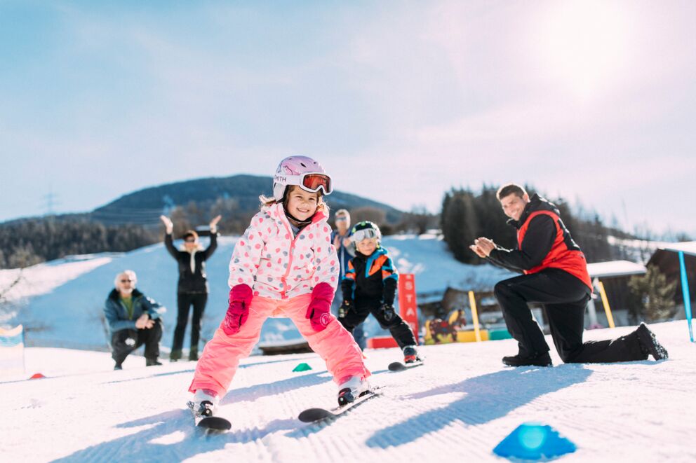 Kinder in Skiausrüstung auf sonniger Piste, von Erwachsenen angefeuert