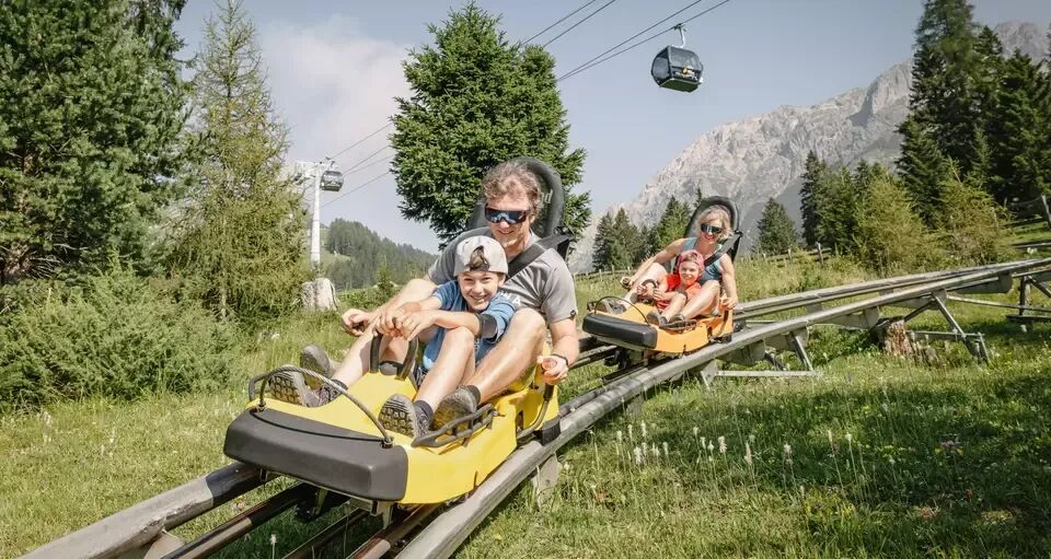 A family rides together on two separate cars on a toboggan run in summer.
