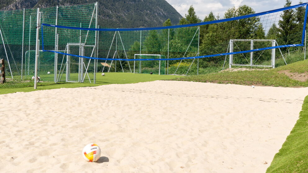 Sandplatz mit Volleyballnetz vor majestätischer Bergkulisse und wolkenverhangenem Himmel.