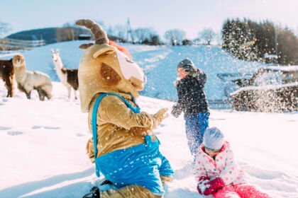 Kinder spielen im Schnee mit einem Maskottchen, Lamas im Hintergrund beobachten.