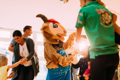 Fröhliche Kinder tanzen mit einem Maskottchen in einem hellen, einladenden Raum.