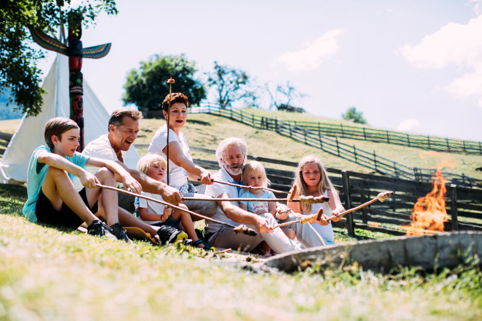 Family grilling sausages and bread together over an open fire outdoors.
