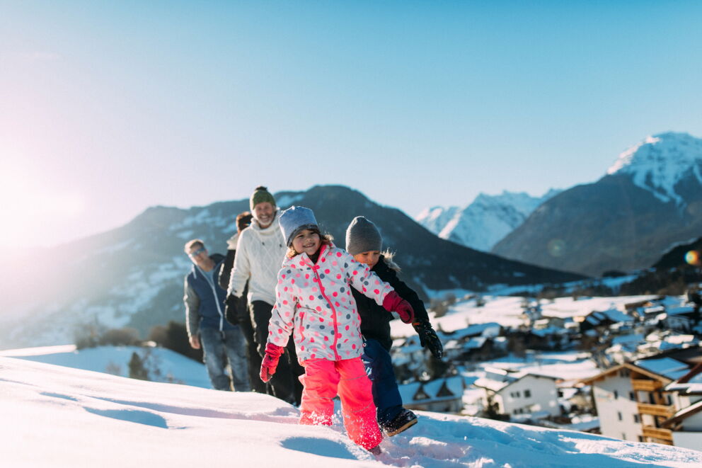 Familie geht lachend im Schnee spazieren, Berge im Hintergrund, sonniger Tag.