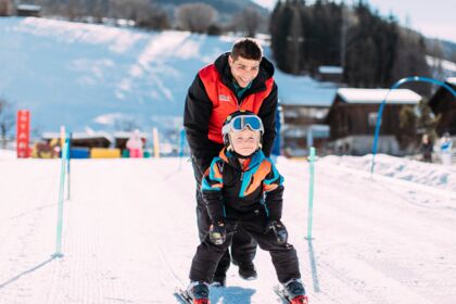 Child learning to ski with the help of an adult on a snow-covered slope.