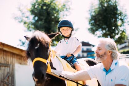 Ein Kind auf einem Pony, begleitet von einem Erwachsenen, bei sonnigem Wetter.