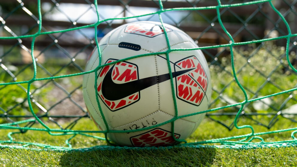 Ein Fußball liegt im Netz auf einem sonnigen, grünen Spielfeld.