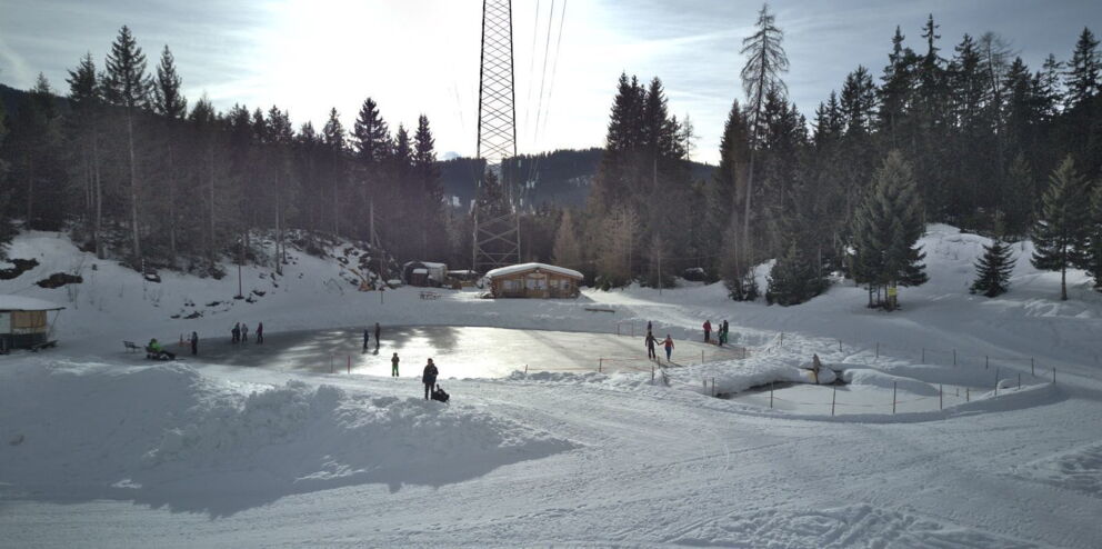 Eislaufplatz mit Kindern umgeben von verschneiten Bäumen und Bergen im Hintergrund.