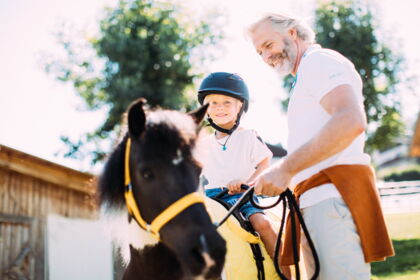 Kind auf Pony mit Helm, begleitet von lächelndem Erwachsenen bei sonnigem Wetter.