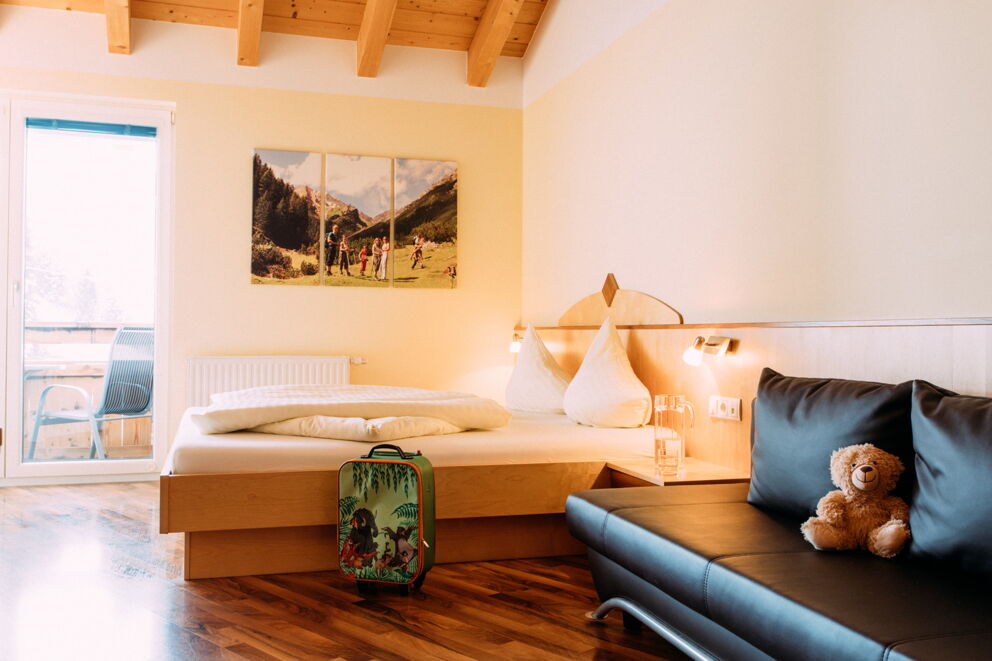 Doppelbett mit Kissen, Couch, Koffer und Teddybär im gemütlichen Zimmer.