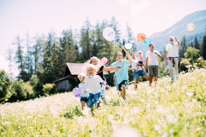 Kinder mit Ballons laufen über sommerliche Wiese, Eltern im Hintergrund freuen sich.