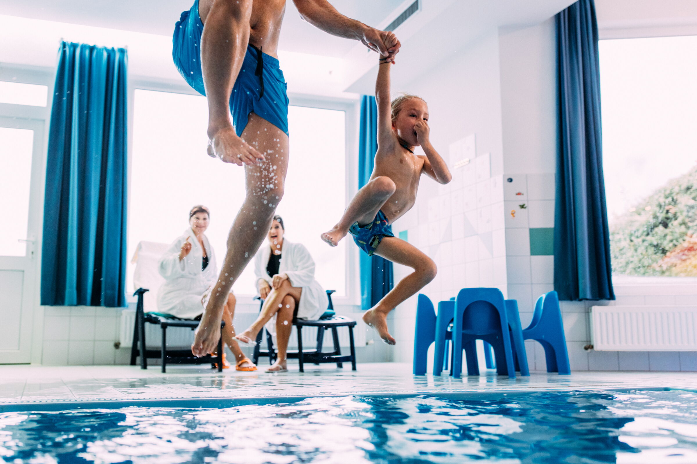 Father and child jump into the pool together while two women look on.