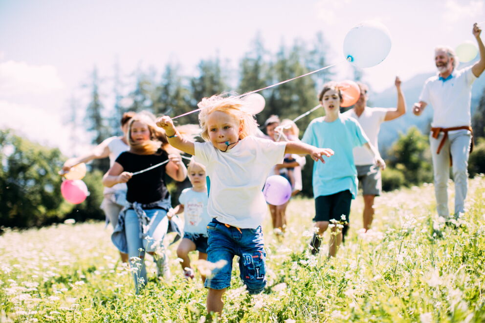 Kinder und Erwachsene laufen mit Ballons auf einer sonnigen Wiese.