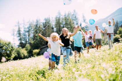 Kinder rennen mit bunten Ballons durch eine blühende Wiese in den Bergen.