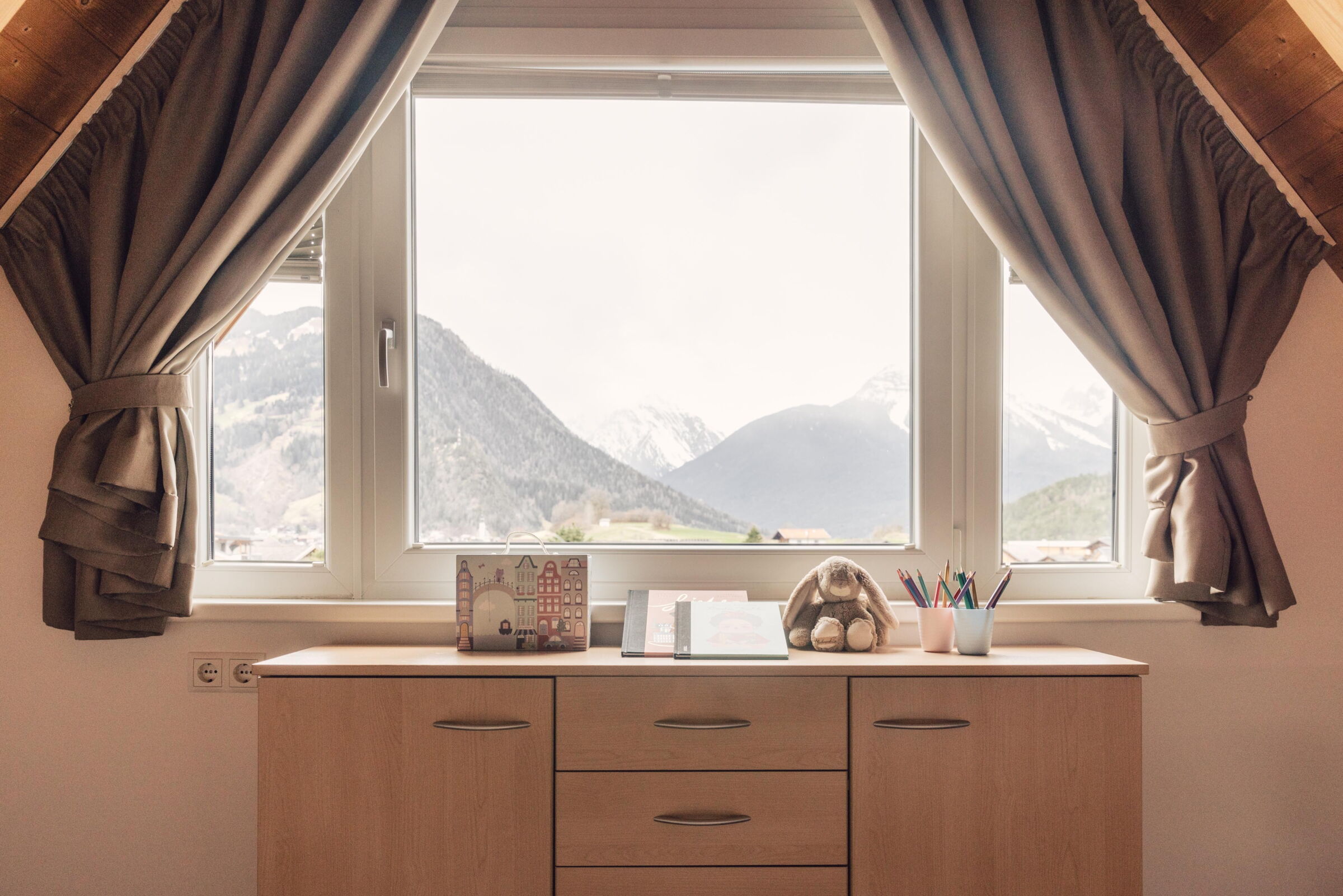 Fenster mit Vorhängen, Holzkommode, Spielzeug, Malbücher und Bergblick im Hintergrund.
