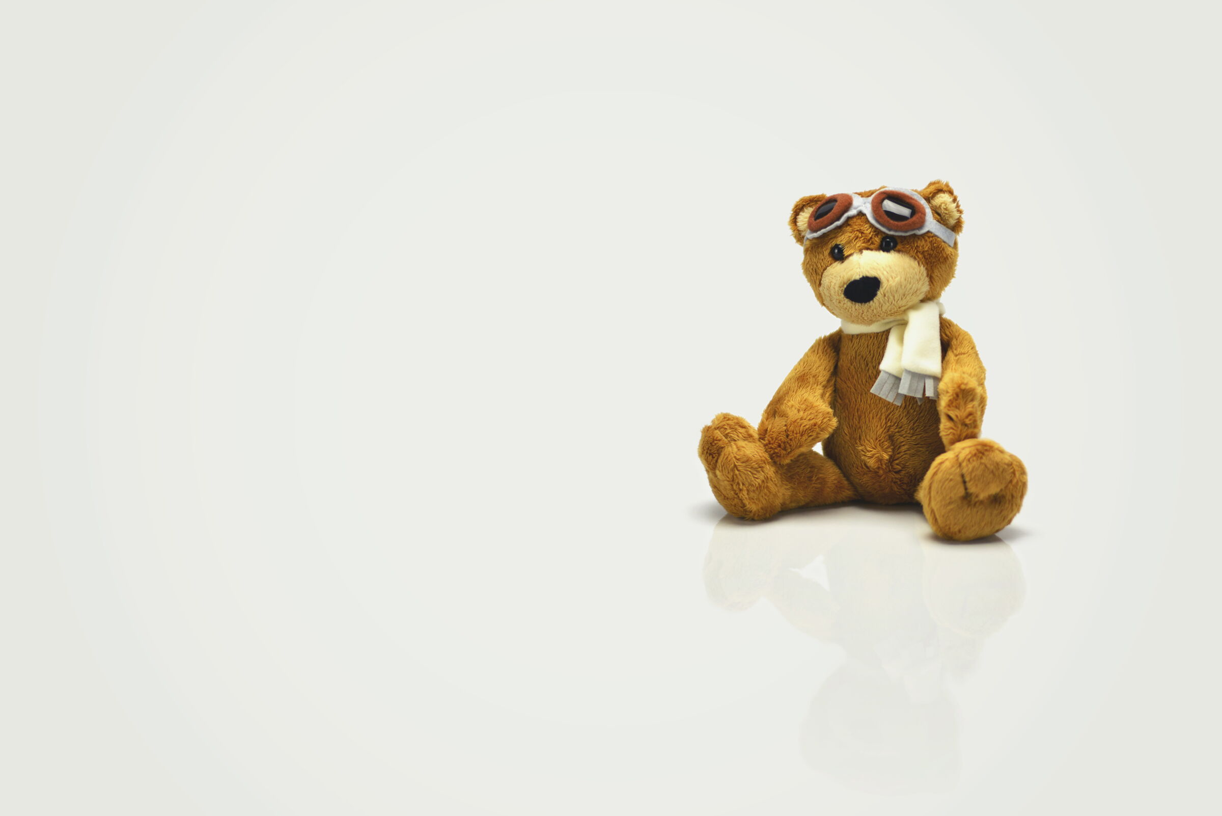 Ein brauner Teddybär trägt Schal und Fliegerbrille, sitzt auf hellem Hintergrund.