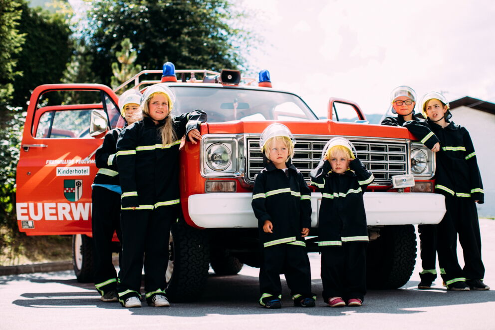 Kinder in Feuerwehrkleidung posieren fröhlich vor einem Feuerwehrwagen im Freien.