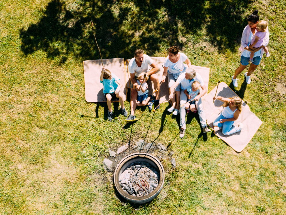 Familien sitzen auf Decken um ein Lagerfeuer, genießen gemeinsam die Natur.
