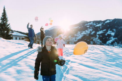 Kinder im Schnee mit bunten Ballons und Bergpanorama im Hintergrund.
