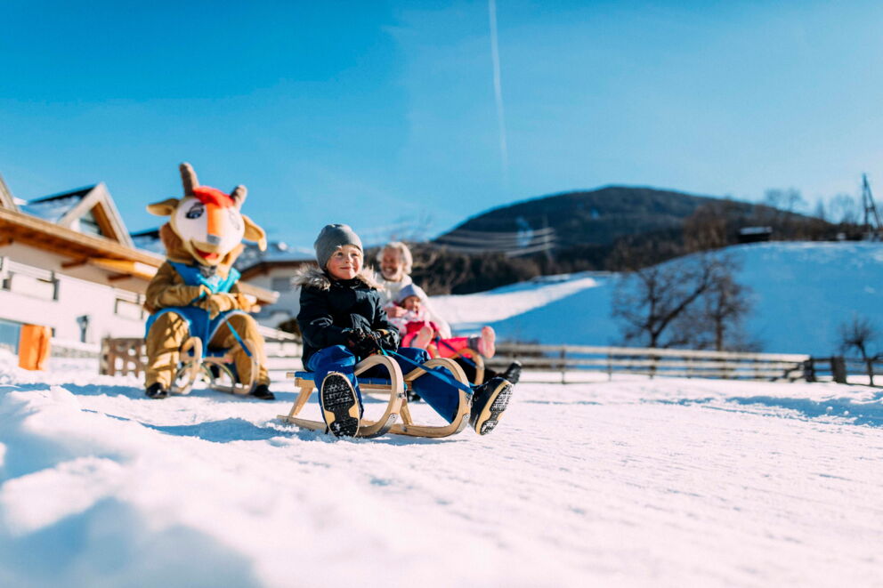 Kinder rodeln im Schnee mit Maskottchen im Hintergrund, sonniger Wintertag.