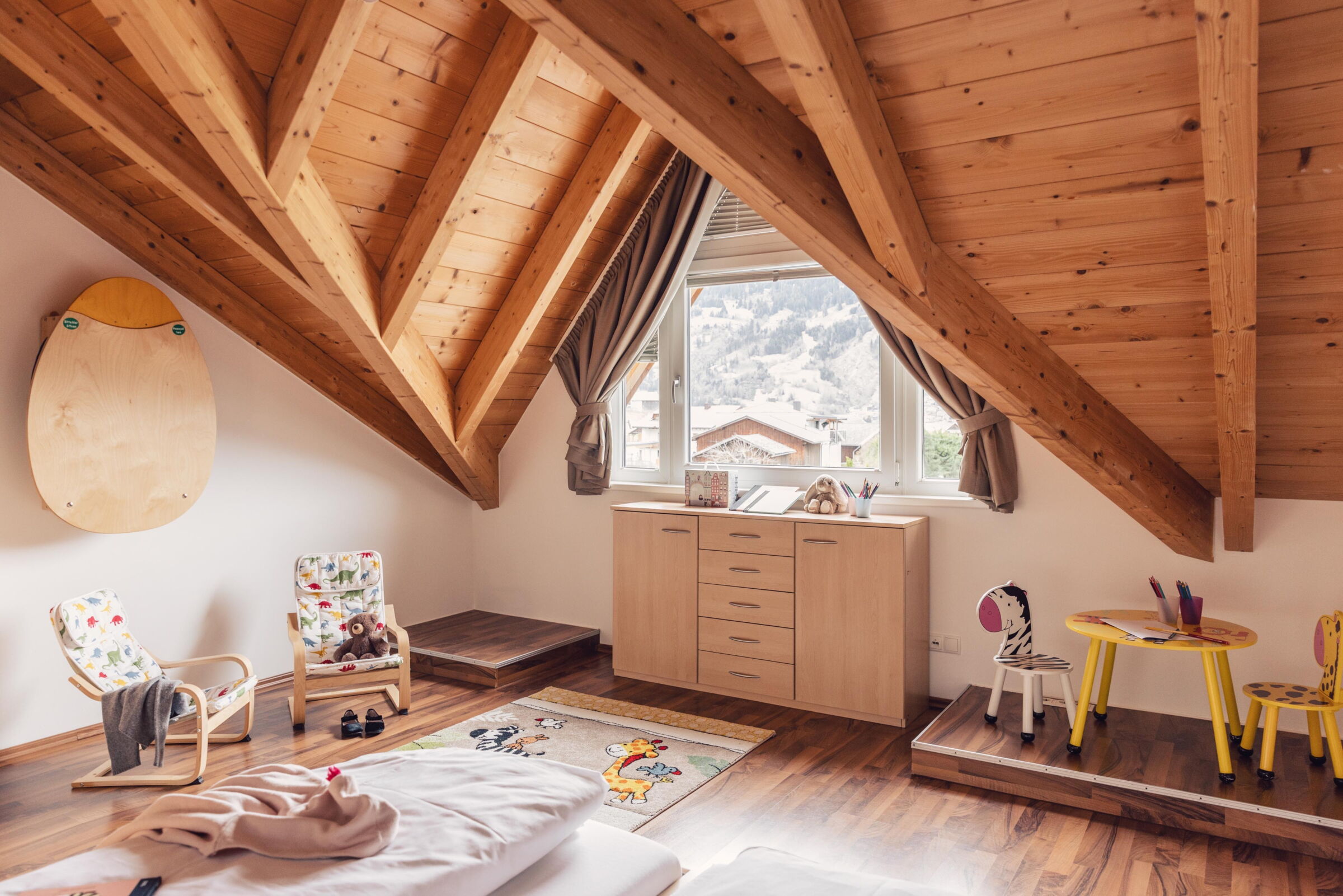 Helles Dachzimmer mit Holzbalken, Kinderstühlen und Bergblick durch das Fenster.