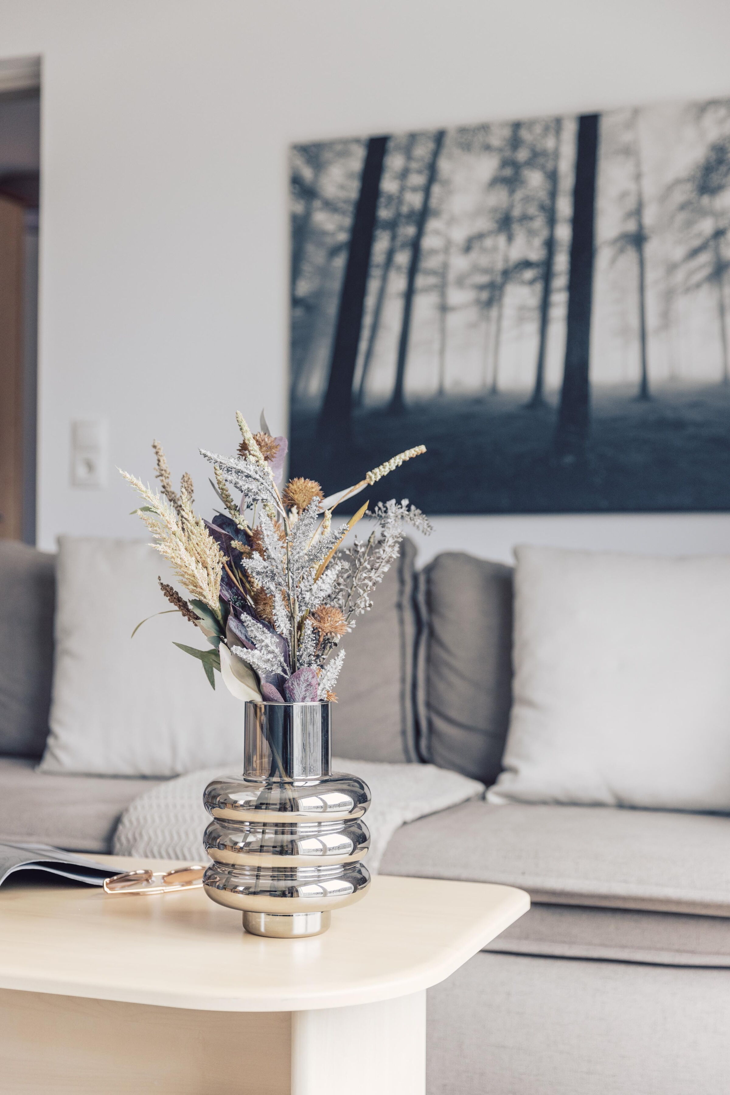 Vase with dried flowers on a wooden table in front of a cozy sofa and forest photography.