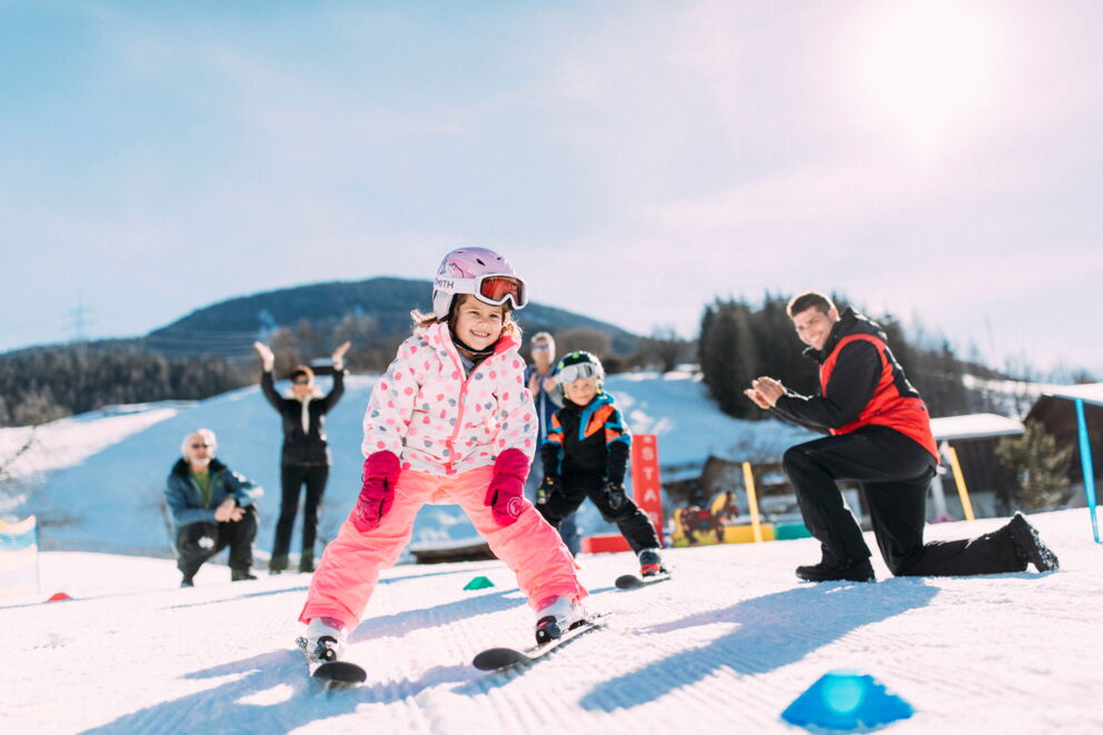 Kinder in Skiausrüstung auf sonniger Piste, von Erwachsenen angefeuert