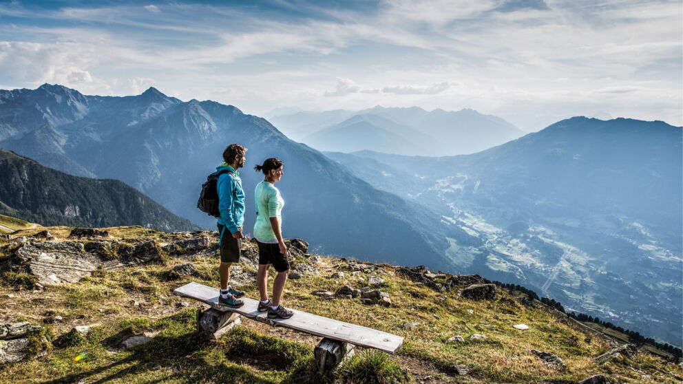 Zwei Wanderer stehen auf einer Bank und blicken auf das Tal in Tirol.