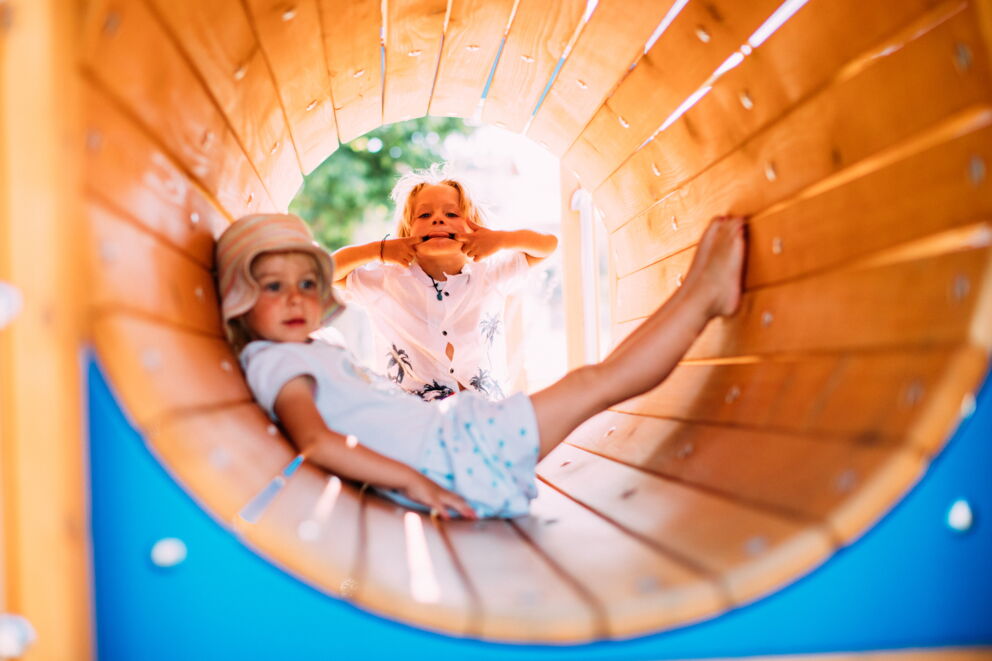 Zwei Kinder spielen lachend in einem hölzernen Tunnel auf einem Spielplatz.