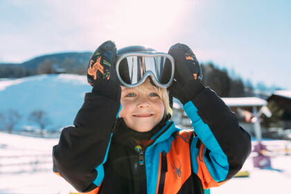 Kind in bunter Winterkleidung mit Skibrille, lächelnd im Sonnenschein.
