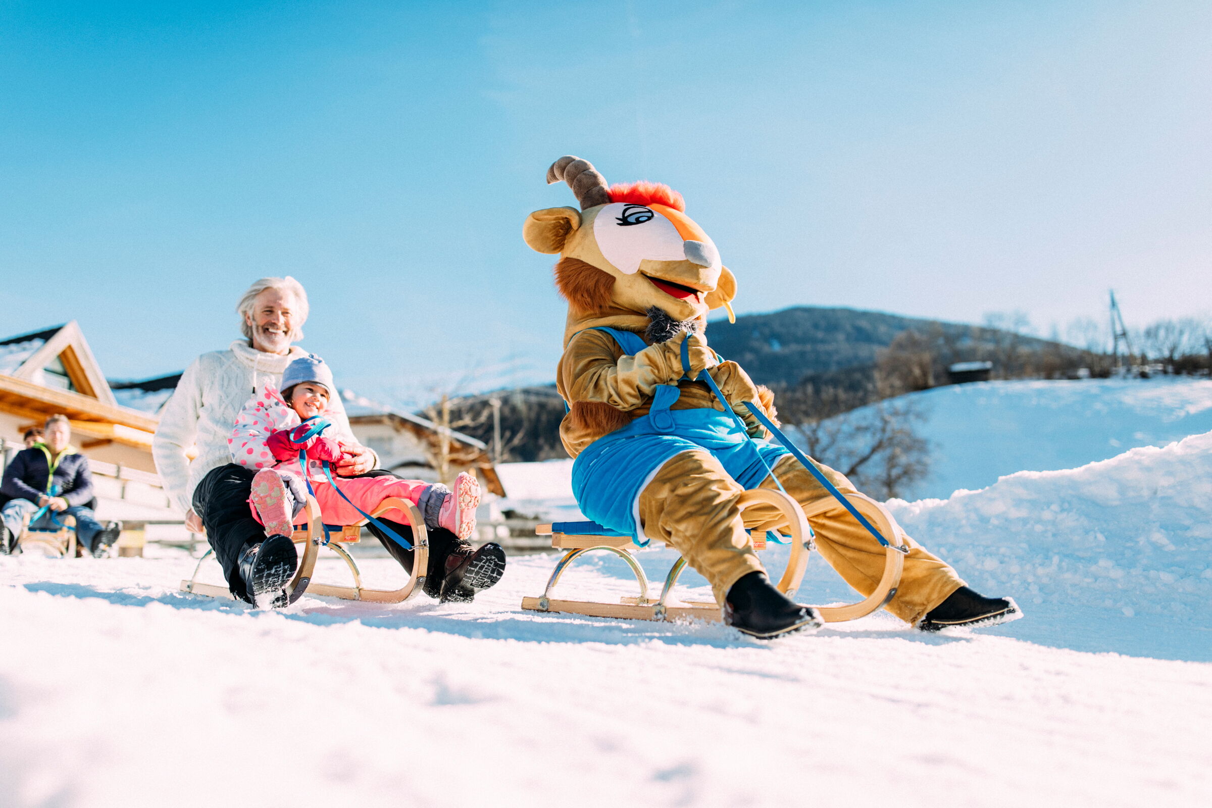 Kind und Erwachsene rodeln fröhlich im Schnee mit Maskottchen im Vordergrund.