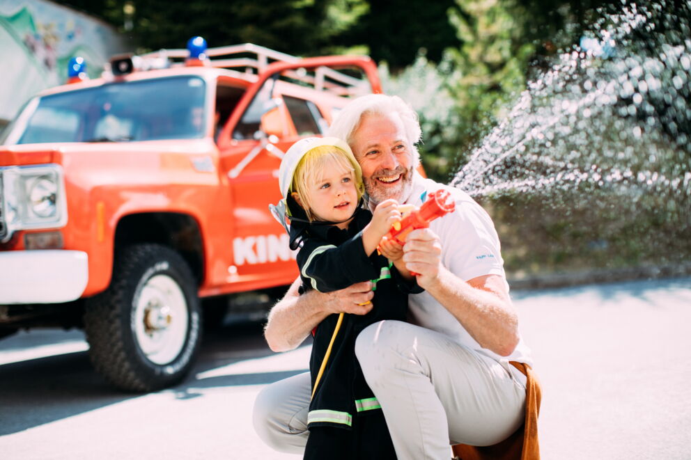 Kind mit Feuerwehrhelm spritzt Wasser, begleitet von einem älteren Mann.