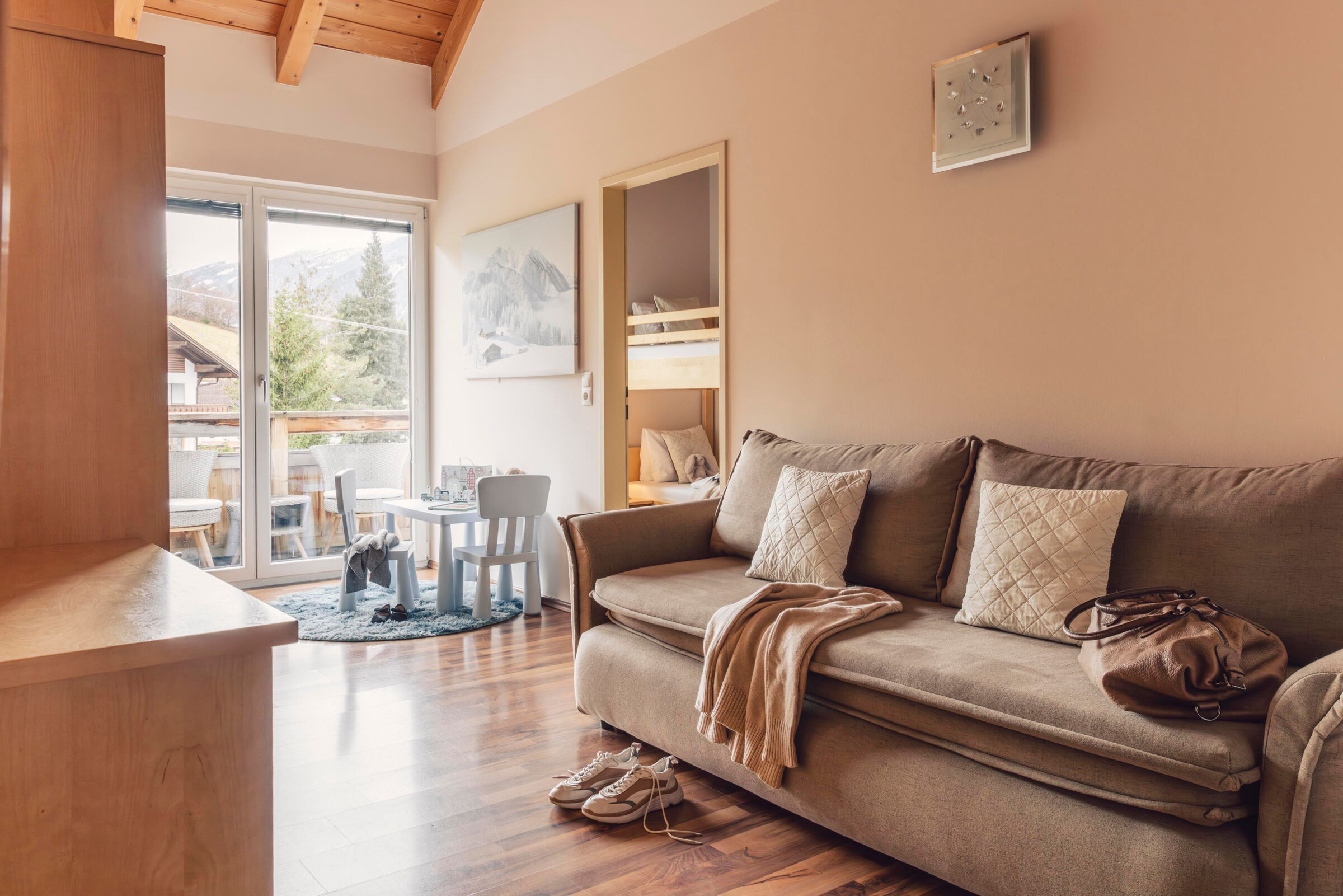 Helles Zimmer mit Holzbalken, Sofa, kleinem Kindertisch und Balkon mit Ausblick.