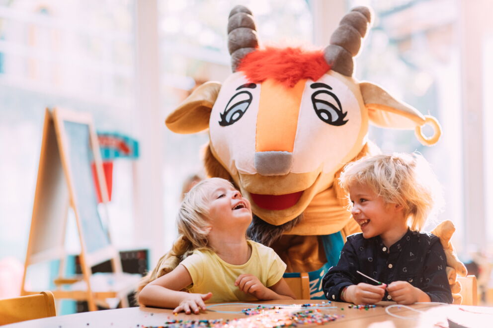Zwei lachende Kinder mit einem Maskottchen in einem hellen Raum.