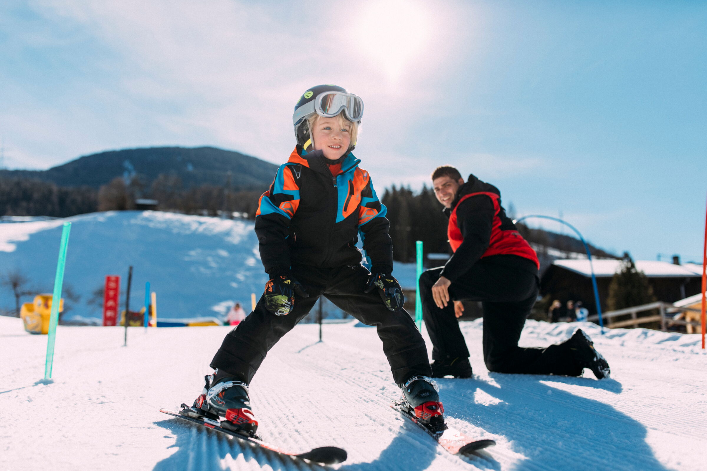 Child learning to ski, supervised by an adult in a sunny winter wonderland.