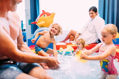 Kinder spielen im Wasser mit buntem Spielzeug, Eltern beobachten lächelnd.