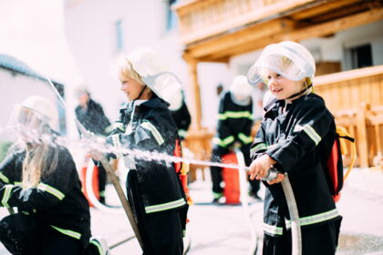 Kinder in Feuerwehrkostümen spielen mit Wasserschläuchen im Freien.
