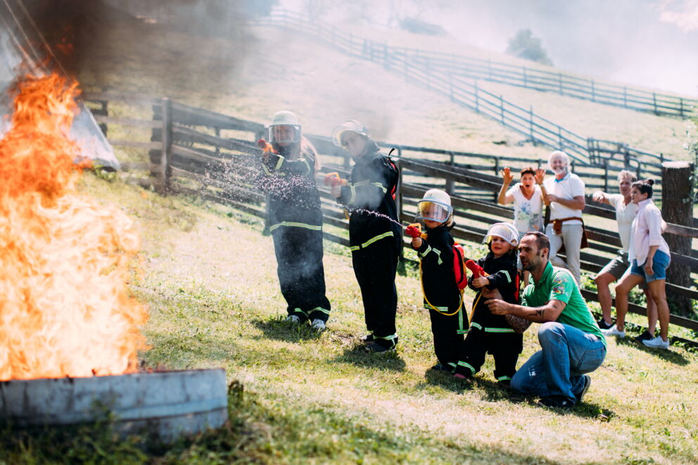 Kinder in Feuerwehranzügen löschen Feuer im Freien, Erwachsene beobachten begeistert.