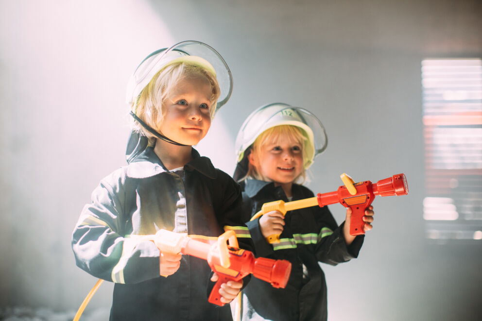 Zwei Kinder in Feuerwehrkostümen mit Spielzeugschläuchen im Lichtstrahl.