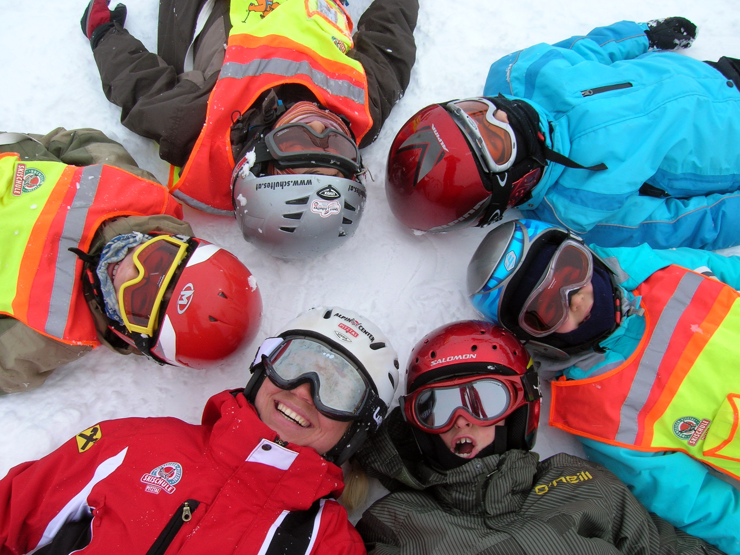 Kindergruppe liegt mit Helmen und Skianzug im Schnee Kopf an Kopf im Kreis
